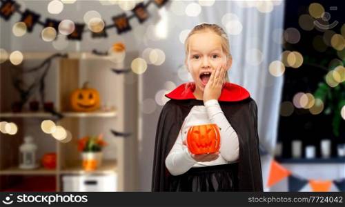 halloween, holiday and childhood concept - happy girl in black dracula cape or costume with jack-o-lantern pumpkin over decorated home room and lights background. girl in halloween costume of dracula with pumpkin