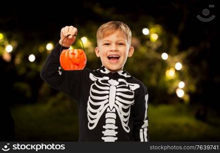 halloween, holiday and childhood concept - happy laughing boy in black costume with skeleton bones holding jack-o-lantern over night park and garland lights on background. happy boy in halloween costume with jack-o-lantern