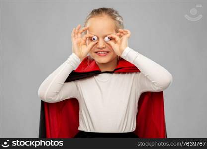halloween, holiday and childhood concept - happy smiling girl in black dracula cape or costume with eyeball candies over grey background. girl in halloween costume of dracula with eyeballs