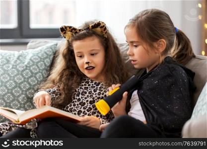 halloween, holiday and childhood concept - little girls in party costumes with flashlight reading book at home. girls in halloween costumes reading book at home