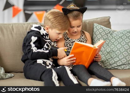 halloween, holiday and childhood concept - little kids in costumes with flashlight reading book at home. kids in halloween costumes reading book at home