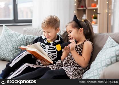 halloween, holiday and childhood concept - little kids in party costumes with flashlight reading book at home. kids in halloween costumes reading book at home