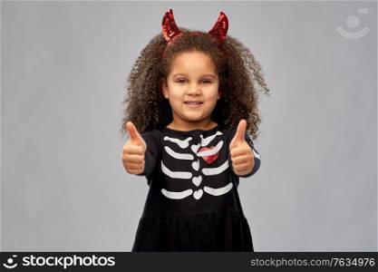 halloween, holiday and childhood concept - smiling african american girl in black costume dress and red devil&rsquo;s horns over grey background. girl in black dress and devil&rsquo;s horns on halloween