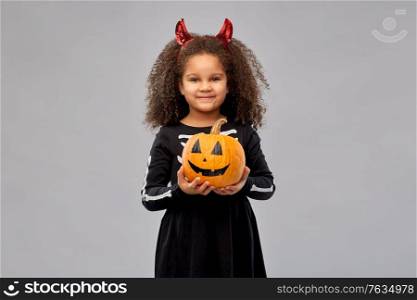 halloween, holiday and childhood concept - smiling african american girl in black costume dress with skeleton bones and red devil&rsquo;s horns holding jack-o-lantern pumpkin over grey background. girl in halloween costume with jack-o-lantern