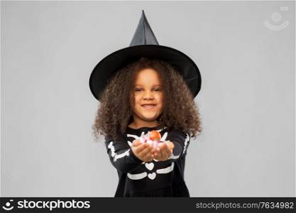halloween, holiday and childhood concept - smiling african american girl in black costume and witch hat with candies trick-or-treating over grey background. girl with candies trick-or-treating on halloween