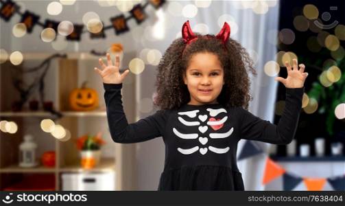 halloween, holiday and childhood concept - smiling african american girl in black costume dress and red devil&rsquo;s horns over decorated home room and festive lights background. girl in black dress and devil&rsquo;s horns on halloween
