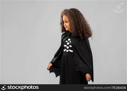 halloween, holiday and childhood concept - smiling african american girl in black dracula costume with bat&rsquo;s cape over grey background. girl in costume of dracula with cape on halloween