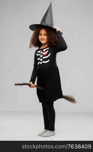 halloween, holiday and childhood concept - smiling african american girl in black costume dress and witch hat with broom over grey background. girl in black witch hat with broom on halloween