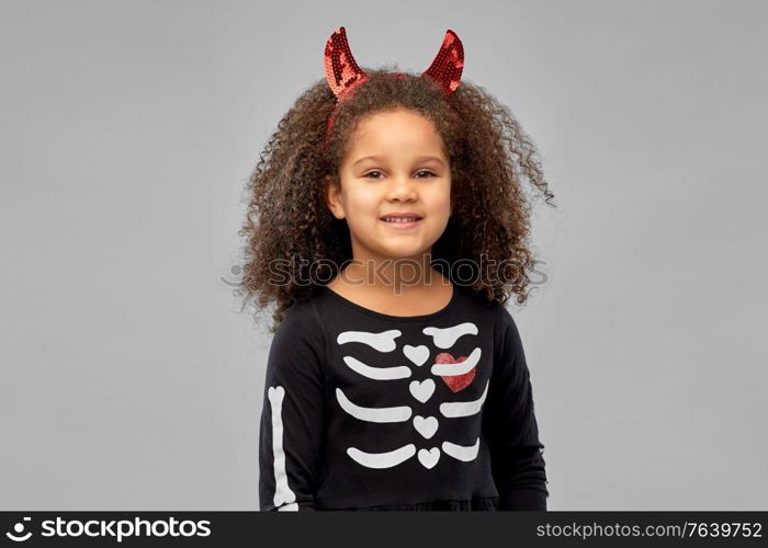 halloween, holiday and childhood concept - smiling african american girl in black costume dress and red devil&rsquo;s horns over grey background. girl in black dress and devil&rsquo;s horns on halloween