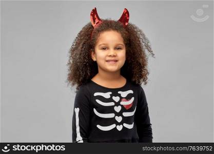 halloween, holiday and childhood concept - smiling african american girl in black costume dress and red devil&rsquo;s horns over grey background. girl in black dress and devil&rsquo;s horns on halloween