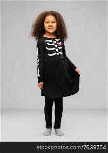 halloween, holiday and childhood concept - smiling african american girl in black costume dress with skeleton bones over grey background. girl in black halloween dress with skeleton bones