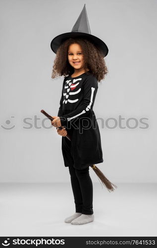 halloween, holiday and childhood concept - smiling african american girl in black costume dress and witch hat with broom over grey background. girl in black witch hat with broom on halloween