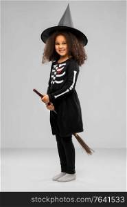 halloween, holiday and childhood concept - smiling african american girl in black costume dress and witch hat with broom over grey background. girl in black witch hat with broom on halloween