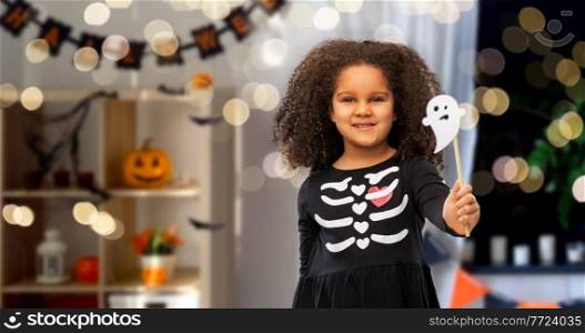 halloween, holiday and childhood concept - smiling african american girl in black costume dress with skeleton bones and ghost over decorated home room and lights background. girl in black halloween dress with skeleton bones