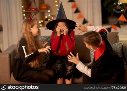 halloween, holiday and childhood concept - smiling boy and girls in costumes playing and scaring each other at home at night. kids in halloween costumes playing at home