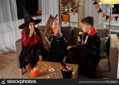 halloween, holiday and childhood concept - smiling boy and girls in party costumes playing with flashlight and scaring each other at home. kids in halloween costumes playing at home