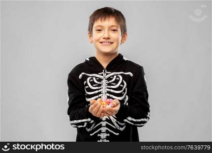 halloween, holiday and childhood concept - smiling boy in black costume of skeleton with candies over grey background. boy in halloween costume of skeleton with candies