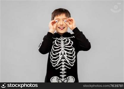 halloween, holiday and childhood concept - smiling boy in black costume of skeleton with eyeball candies over grey background. boy in halloween costume of skeleton with eyeballs