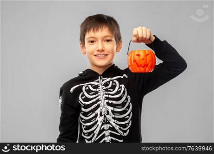 halloween, holiday and childhood concept - smiling boy in black costume of skeleton with jack-o-lantern pumpkin over grey background. boy in halloween costume with jack-o-lantern