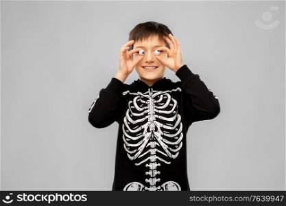 halloween, holiday and childhood concept - smiling boy in black costume of skeleton with eyeball candies over grey background. boy in halloween costume of skeleton with eyeballs