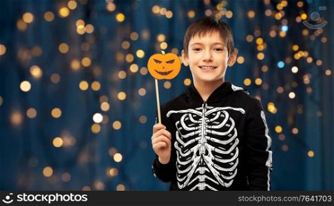 halloween, holiday and childhood concept - smiling boy in black costume of skeleton with jack-o-lantern pumpkin party prop over festive lights on background. boy in halloween costume of skeleton with pumpkin