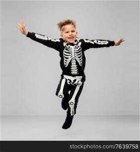 halloween, holiday and childhood concept - smiling boy in black costume with skeleton bones jumping over grey background. happy boy in halloween costume of skeleton jumping