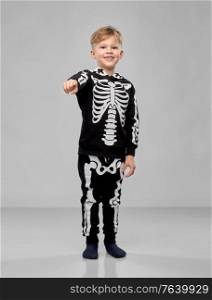 halloween, holiday and childhood concept - smiling boy in black costume with skeleton bones pointing to camera over grey background. boy in costume of skeleton pointing to camera