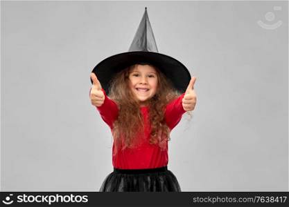 halloween, holiday and childhood concept - smiling girl in costume and witch hat showing thumbs up over grey background. happy girl in black witch hat on halloween