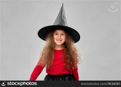 halloween, holiday and childhood concept - smiling girl in costume and witch hat over grey background. happy girl in black witch hat on halloween
