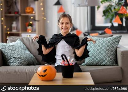 halloween, holiday and childhood concept - smiling girl in costume with bat cape at home. girl in halloween costume with bat cape at home