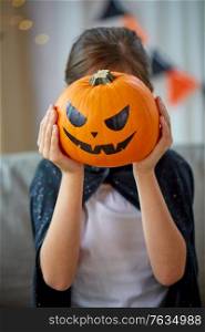 halloween, holiday and childhood concept - smiling girl in costume with jack-o-lantern at home. girl in halloween costume with pumpkin at home