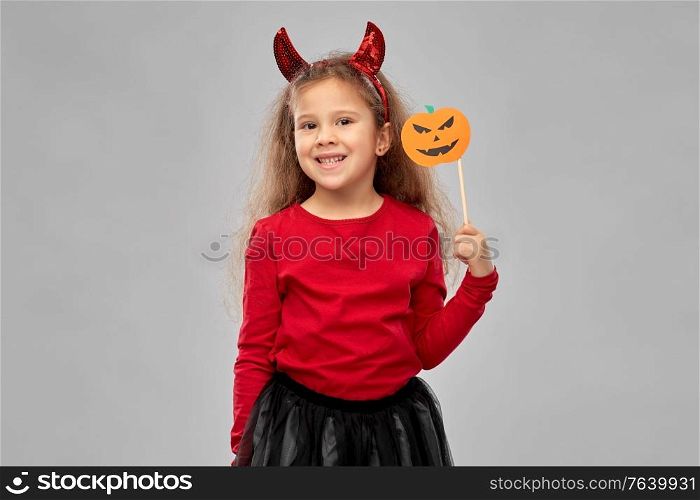 halloween, holiday and childhood concept - smiling girl in costume with red devil&rsquo;s horns holding jack-o-lantern pumpkin party accessory over grey background. girl in halloween costume with jack-o-lantern
