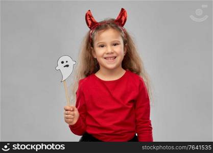 halloween, holiday and childhood concept - smiling girl in costume with red devil&rsquo;s horns holding ghost party accessory over grey background. girl in halloween costume with ghost party prop