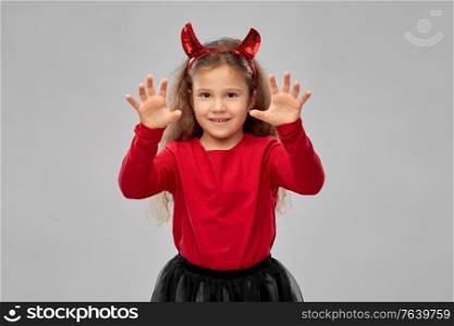 halloween, holiday and childhood concept - smiling girl in party costume with red devil&rsquo;s horns making spooky gestures over grey background. girl costume with devil&rsquo;s horns on halloween