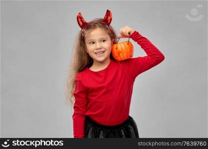 halloween, holiday and childhood concept - smiling girl in party costume with red devil&rsquo;s horns holding jack-o-lantern pumpkin over grey background. girl in halloween costume with jack-o-lantern