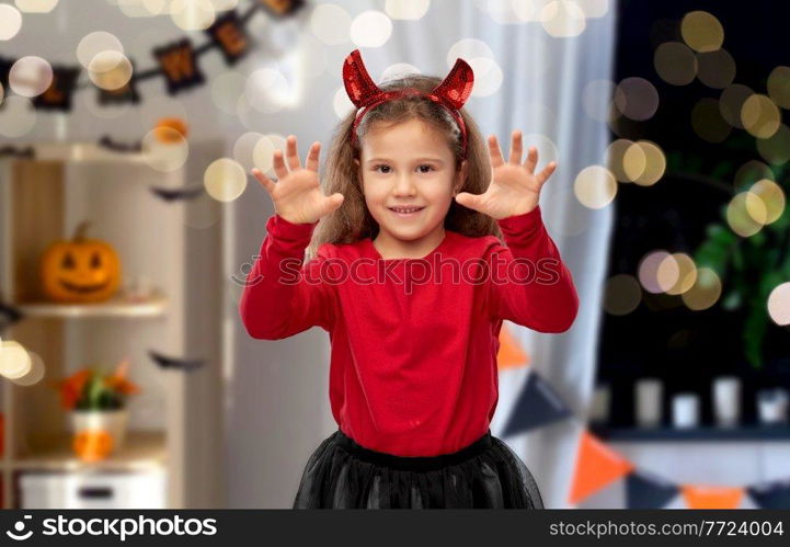 halloween, holiday and childhood concept - smiling girl in party costume with red devil’s horns making spooky gestures over decorated home room and lights background. girl costume with devil’s horns on halloween