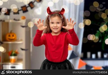 halloween, holiday and childhood concept - smiling girl in party costume with red devil’s horns making spooky gestures over decorated home room and lights background. girl costume with devil’s horns on halloween