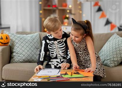 halloween, holiday and childhood concept - smiling little boy and girl in costumes doing crafts at home. kids in halloween costumes doing crafts at home