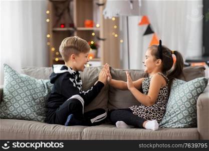 halloween, holiday and childhood concept - smiling little boy and girl in costumes playing clapping game at home. kids in halloween costumes playing game at home