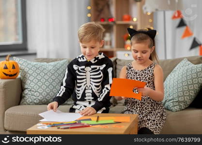 halloween, holiday and childhood concept - smiling little boy and girl in costumes doing crafts at home. kids in halloween costumes doing crafts at home