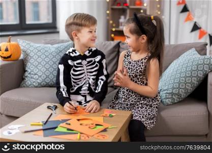 halloween, holiday and childhood concept - smiling little boy and girl in party costumes doing crafts at home. kids in halloween costumes doing crafts at home