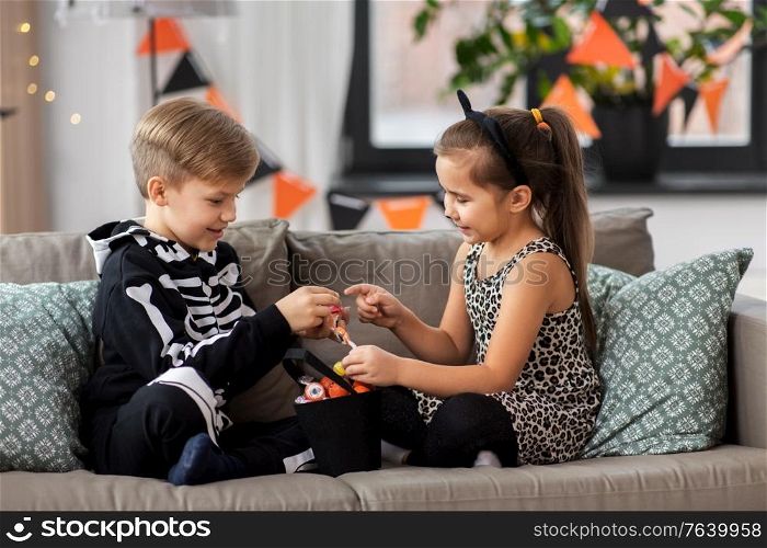 halloween, holiday and childhood concept - smiling little boy and girl in costumes with candies sitting on sofa at home. kids in halloween costumes with candies at home