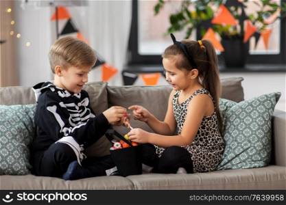 halloween, holiday and childhood concept - smiling little boy and girl in costumes with candies sitting on sofa at home. kids in halloween costumes with candies at home