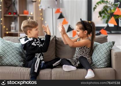 halloween, holiday and childhood concept - smiling little boy and girl in costumes playing clapping game at home. kids in halloween costumes playing game at home