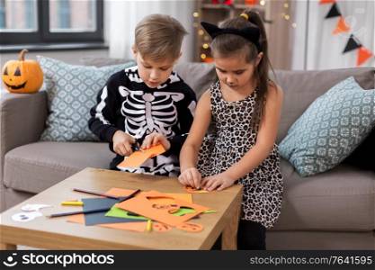 halloween, holiday and childhood concept - smiling little boy and girl in party costumes doing crafts at home. kids in halloween costumes doing crafts at home