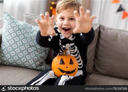 halloween, holiday and childhood concept - smiling little boy in party costume of skeleton with jack-o-lantern pumpkin sitting on sofa and having fun at home. happy boy in halloween costume of skeleton at home