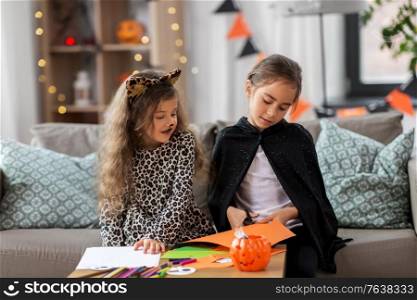 halloween, holiday and childhood concept - smiling little girls in costumes doing crafts at home. girls in halloween costumes doing crafts at home