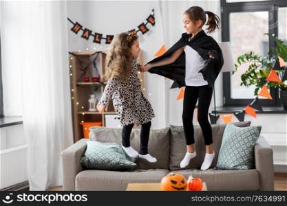 halloween, holiday and childhood concept - smiling little girls in costumes jumping on sofa at home. girls in halloween costumes jumping on sofa