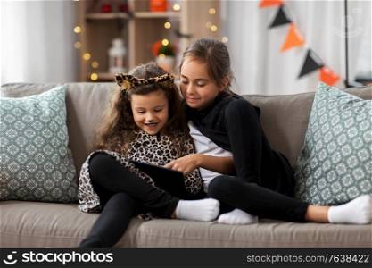 halloween, holiday and childhood concept - smiling little girls in costumes with tablet pc computer at home. girls in halloween costumes with tablet pc at home
