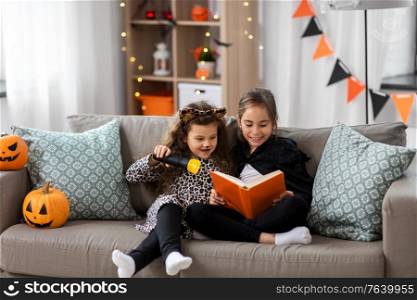 halloween, holiday and childhood concept - smiling little girls in costumes with flashlight reading book at home. girls in halloween costumes reading book at home